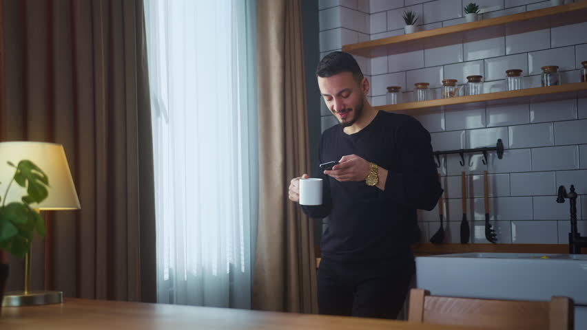 Smiling man standing in modern kitchen having morning coffee, holding cell phone scrolling news feed, checking emails, social media on smartphone
