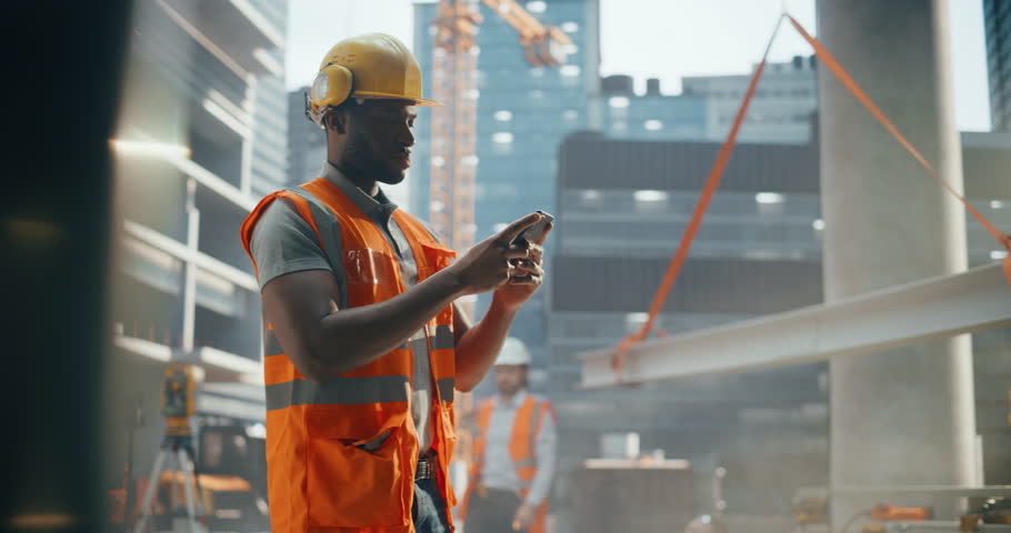 Portrait of Handsome African Builder, Worker, Craftsman Wearing a Hard Hat and a Vest while Standing at a Commercial Building Construction Site and Using a Smartphone to Browse Through Social Media Royalty-Free Stock Footage #3434913167