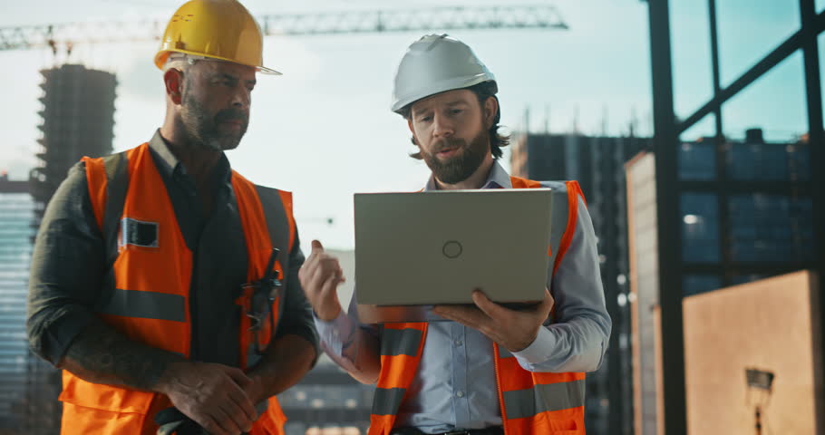 Construction and Site Managers Having a Meeting at a Construction Site with Crane Operator Working in the Background. Specialists Using Laptop Computer to Review the Technical Drawings Royalty-Free Stock Footage #3434918349