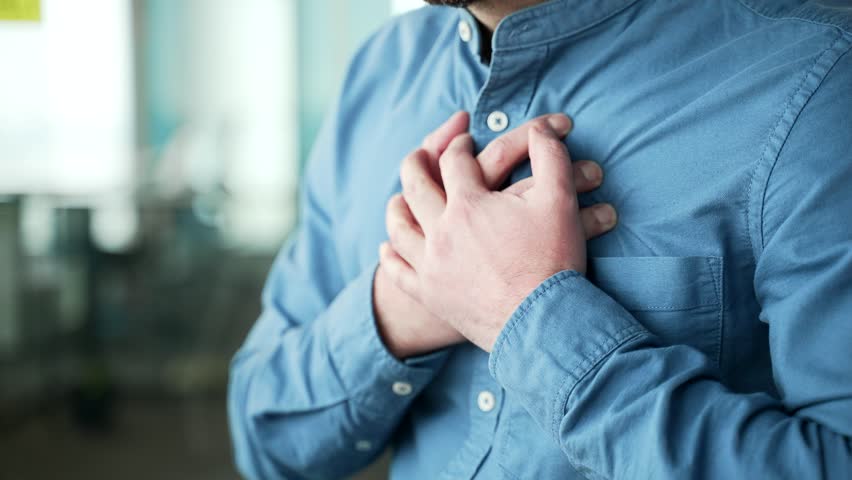Close up. Male hands holding massaging chest. Mature businessman in a blue shirt has a heart attack while sitting at a workplace in a business office. Sick man is breathing deeply, needs medical help