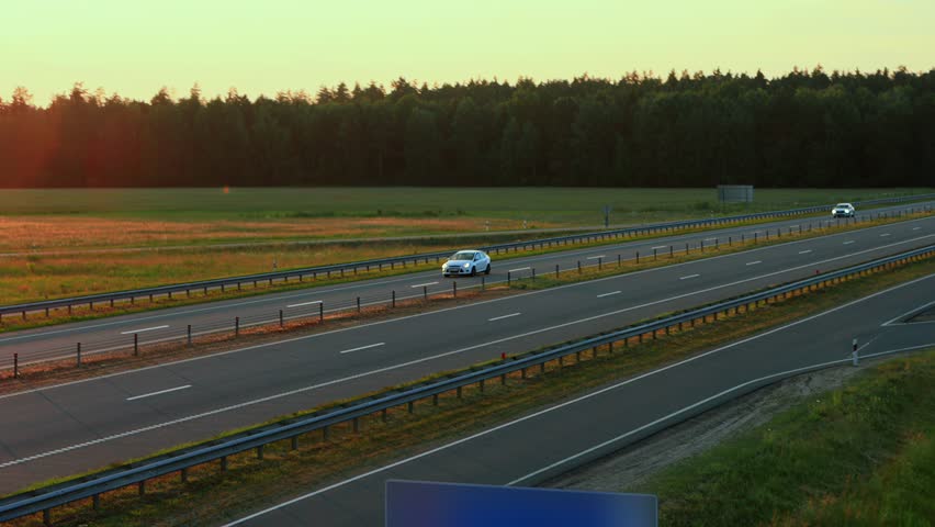 White sedan car driving fast on the highway at sunset, two lane roadway. Traffic barrier guard rail along throughway. Compliance with speed limits on motorway countryside, out of town Royalty-Free Stock Footage #3434941261