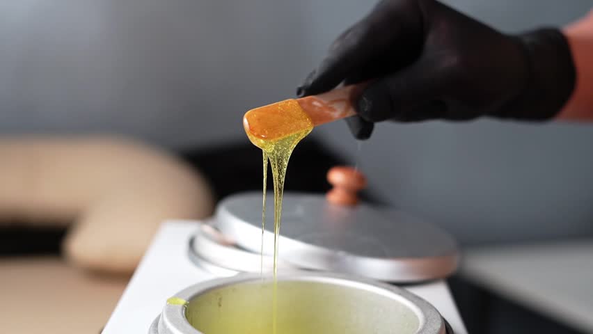 Hot gold wax used for hair removal. Close-up of depilation master hands in black protective gloves holds spatula with melted wax. Device for melting wax