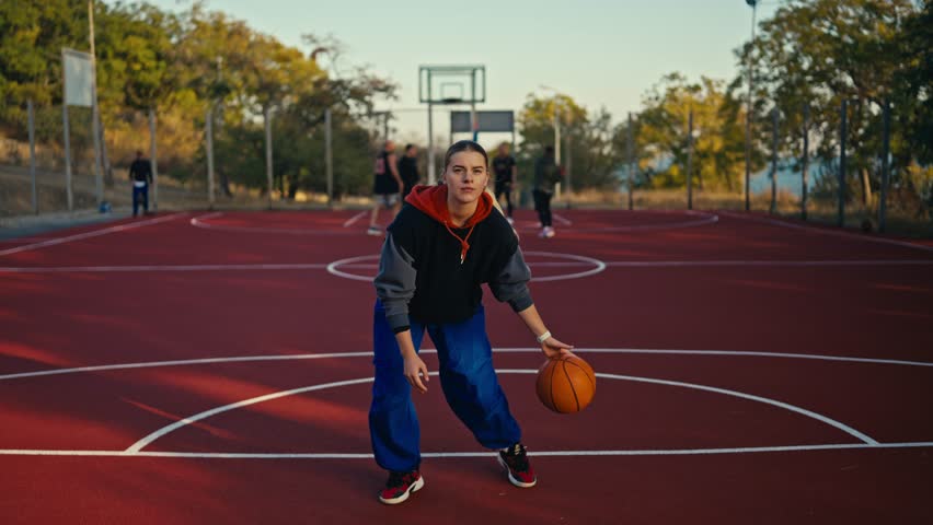 Portrait of a blonde girl in sportswear and blue pants who bounces an orange basketball ball off the floor and maneuvers with it and then throws it into the hoop on a red basketball court on the - Powered by Shutterstock - Get 15% off with code: PIKWIZARD15