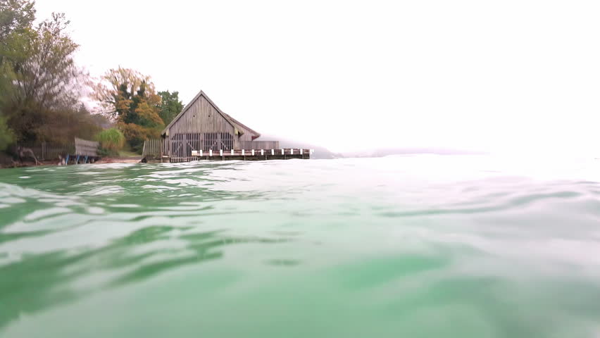 Wooden hut on lakeside with mist covered mountains in distance. Waves crashing into camera. 