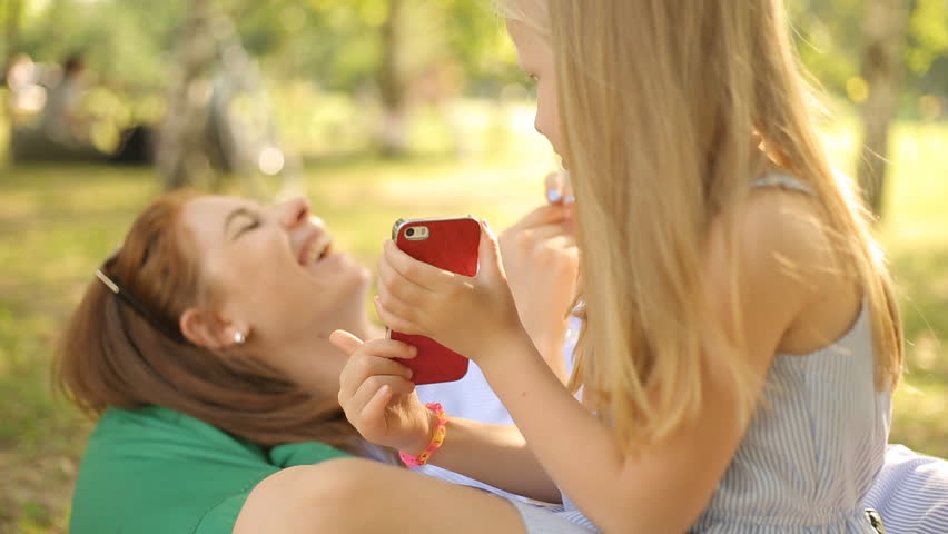 A Little Cute Girl Is Sitting On A Bench On The Street Holding A Smartphone And Typing