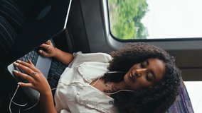Vertical video of young businesswoman commuting sitting by window on moving train passing through city working on laptop and listening to music or podcast on earphones - shot in slow motion