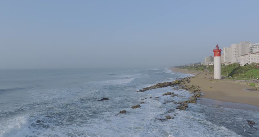 The umhlanga lighthouse on a sunny day with waves crashing against the shore, urban backdrop, aerial view