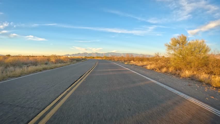 POV - Driving on a asphalt road with some traffic through Sonoran Desert in Arizona; Santa Rita mountains on the horizon; late afternoon sun casing long shadows and illuminating the local flora