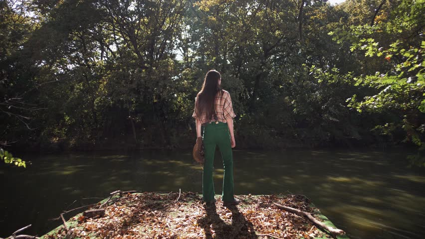 Young teenager with long hair wearing a plaid shirt stands on a dock by a river in the woods, holding a wicker hat in his hands 