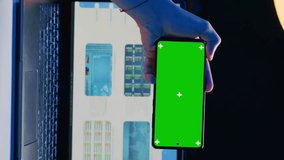 Vertical video Man sitting on chair at home office desk in neon illuminated apartment holding green screen phone while working. Person in living room looking at business graphs on laptop and at mockup