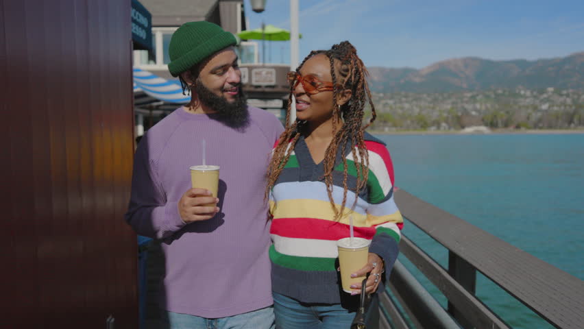 Black young couple enjoys leisure time on a pier with coffee, smiling while overlooking the ocean. The man's beard and hat add to their sense of fun and adventure. Santa Barbara, USA Royalty-Free Stock Footage #3435513007