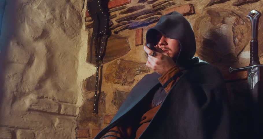 Man smoker hides from falling snow sitting by tavern entrance. Mysterious frozen guest in long hood smokes pipe sitting in pub. Medieval winter fantasy