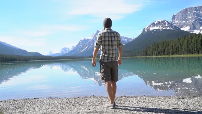Slow motion video of a young man jumping mid-air by a mountain lake in Banff national park, Alberta, Canada. Beautiful Rocky Mountains on the background. People travel success concept