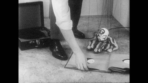 1950s: Marionette sits and watches man put record on phonograph. Man turns on record player. Puppet dances.