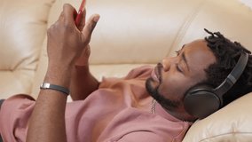 Portrait of african american man in headphones lying on couch browsing phone. Sleepy relaxed guy surfing online on websites social media at home, side view. Entertainment, leisure with mobile phone.