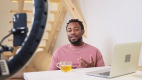 African american man blogger hipster recording video on smartphone with ring lamp sitting at table with cup of tea and laptop. Smiling positive sociable guy vlogger creating content for vlog. - Powered by Shutterstock - Get 15% off with code: PIKWIZARD15