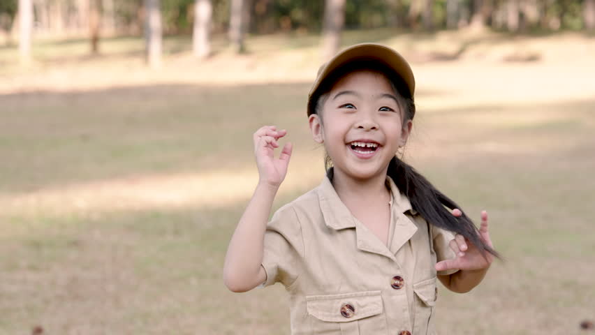 Joyful Asian girl with raised arms, khaki cap and shirt, celebrating in sunlit park. Cheerful child in khaki, arms up, exuding happiness in bright outdoor setting.