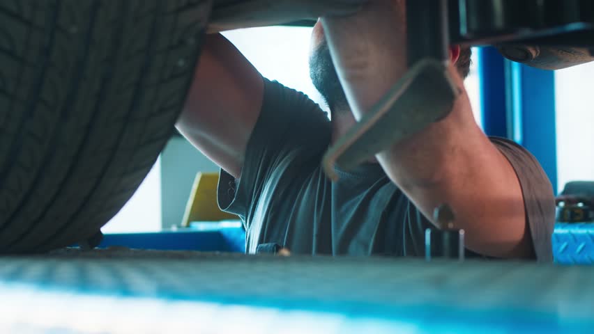 Technician attentively inspects the undercarriage, tools at hand, in the blue-hued ambiance of garage. Royalty-Free Stock Footage #3435856567