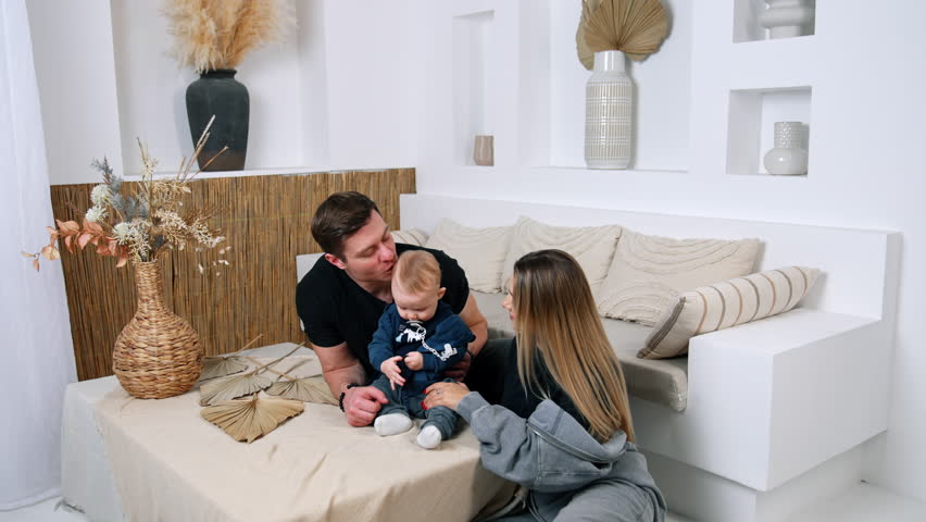 Infant boy with pacifier sits on the table. Parents support their cute child sitting behind.