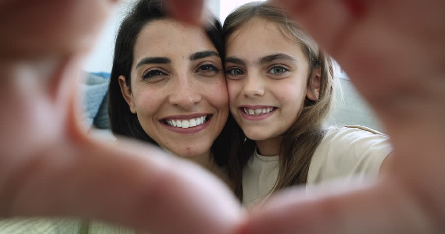 Close up happy mom and adopted daughter take selfie portrait, show love sign, heart gesture, look at camera through joined fingers. Smiling foster mom with little kid girl have fun. Adoption, custody