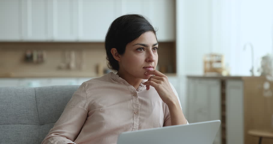 Pensive Indian woman sits on couch at home with laptop, work remotely, do freelance on-line job, makes creative university assignment, typing essay or summary, think over solution, looks thoughtful Royalty-Free Stock Footage #3436199493