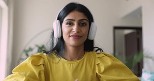 Attractive Indian woman in wireless headphones looking at camera make speech, provide professional consultation to client use videocall application. Webcam view female talk to relatives by video call