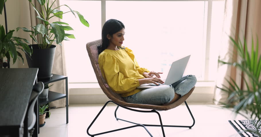 Pretty Indian woman working on laptop seated in modern armchair in living room. Focused female browsing web site, share messages by messenger app or e-mailing, studying online, do remote freelance job Royalty-Free Stock Footage #3436255951