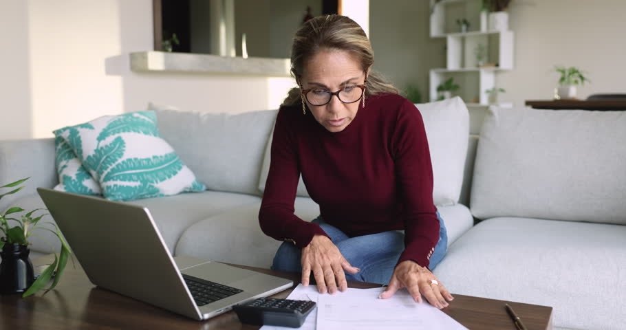 Older Latina woman do accounting job from home, sit on couch at table with laptop, make on-line payments using e-bank app, review spendings and savings, calculates earnings, paying taxes and mortgage Royalty-Free Stock Footage #3436281579