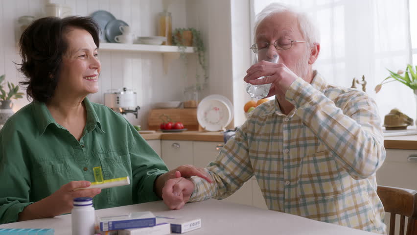 elderly man and woman sit at table in kitchen in morning. takes pills and wash dawn with water. wife supports and cares her husband health. looks kindly and gently smiles, reminding of medications