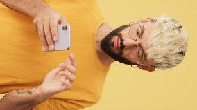 Vertical video, Guy with glasses, dressed in yellow T-shirt, with phone in his hands scrolls, chooses, makes purchases, isolated on yellow background in studio