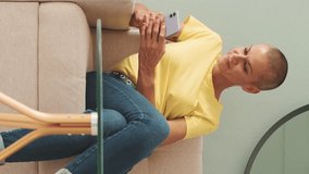 Vertical video, Smiling bald woman writing on social networks with mobile phone while sitting in living room