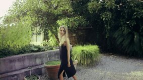 A cheerful blonde woman model girl wearing a black dress walking around in the summer garden towards the camera holding her phone in hand and the old European building in the background, 4K video
