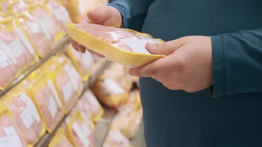 Man chooses package of organic chicken breast fillet in a grocery supermarket standing at the shelves with products. Male looks at the price and expiration date of the product in a shop