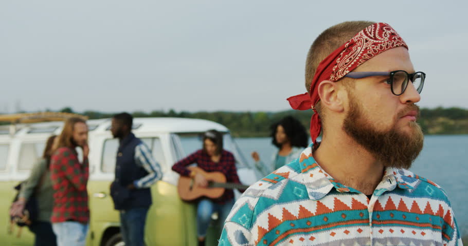 Young male hipster in glasses standing in front of the camera. Group of the mixed races friends on the background near a green van and a lake. Outside. Portrait shot. Close up