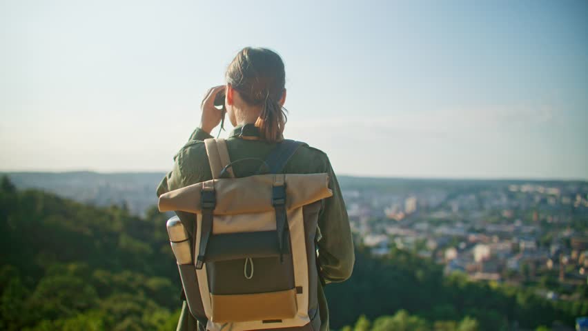 View from behind of Caucasian girl with ponytail and backpack. Young woman observing beautiful landscape in front of her with binoculars. Enjoying wonderful scenery. Active lifestyle.