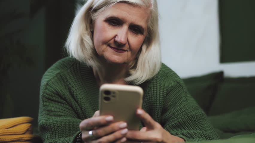 Attractive senior woman playing a game on mobile phone, typing a message on smartphone, chatting online. Retired old grandmother Female holding gadget, using mobile device while lying down on a bed Royalty-Free Stock Footage #3436962537