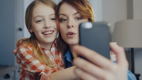 Close up of the smiling mother hugging young daughter and making funny selfies on the smartphone while grimacing. Portrait. Inside