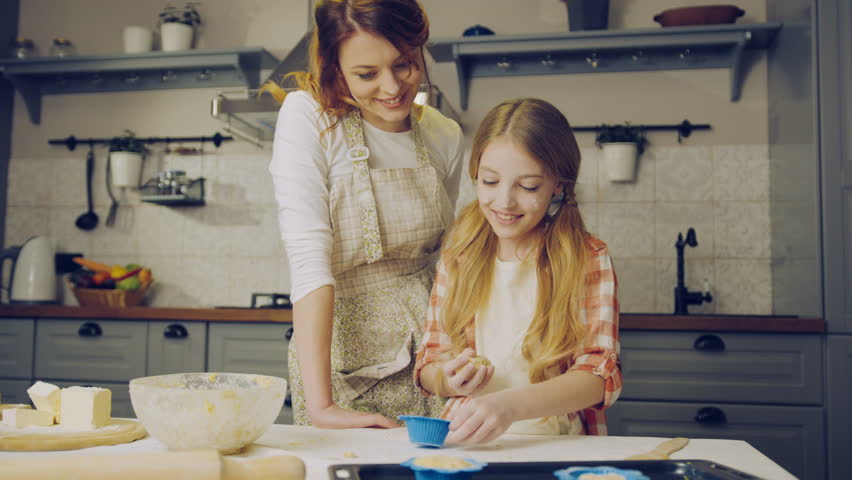 Attractive mother watching her blonde daughter preparing daugh for muffins and putting it in the forms. 