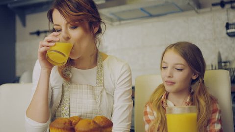 Attractive mother in the apron drinking juice and her cute daughter kissing her in the cheek, than mom and daughter kissing and hugging in the kitchen. Indoors