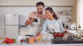 Joyful Indian couples cook or trying new food recipe by watching online videos from Mobile phone at Kitchen - concept of technology, relationship bonding and collaboration