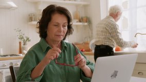 elderly woman sits at table in kitchen with laptop. types on keyboard looks intently in screen, smiles. succeed in using computer and internet, feels pleased and comfortable. old man on background - Powered by Shutterstock - Get 15% off with code: PIKWIZARD15