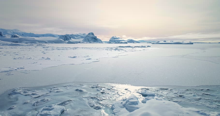 Fly over frozen Antarctic ocean sunset landscape. Snow covered untouched wilderness of South Pole. Desert white land of snow and ice aerial drone shot. Mountains in background. Nature conservation