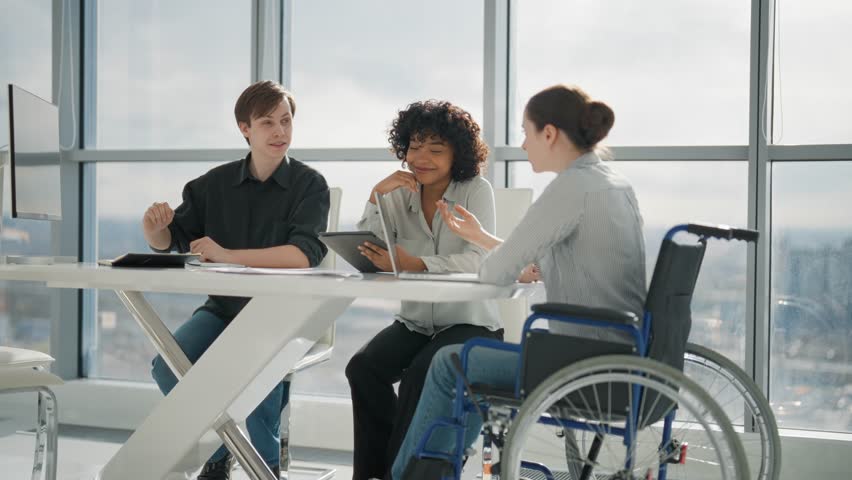 Corporate colleagues working on project in office together sitting at desk. Woman in wheelchair with physical disability on meeting with caucasian man african american female. Teamwork, discuss. Royalty-Free Stock Footage #3437073549