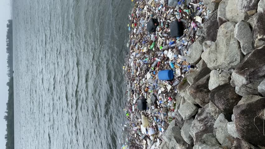 Rocky shoreline facing Johor Straits, polluted with rubbish and plastic waste built up, floating in the sea. Pollution brought by waves to the shore. Vertical video slow pan right. Royalty-Free Stock Footage #3437134825