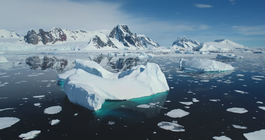 Aerial flight above giant iceberg with melting hole cavity inside. Snow covered glaciers and mountain range in background. Polar winter landscape. Sunny day blue sky. Global warming and climate change