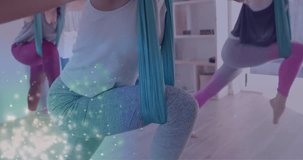 Animation of glowing green particles over happy diverse teenage girls practicing aerial yoga. Yoga, wellbeing, energy, healthy lifestyle, connection and communication, digitally generated video.