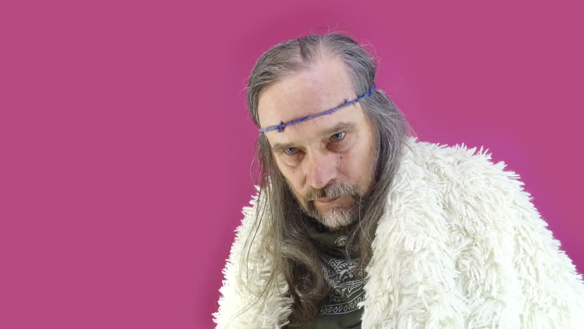 Portrait gray-haired mature Viking with long hair looks menacingly into distance, dressed in white goat fur coat on pink green background