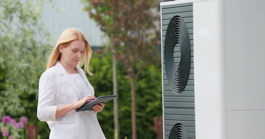 Woman sets up a heat pump near a private house. Uses a tablet