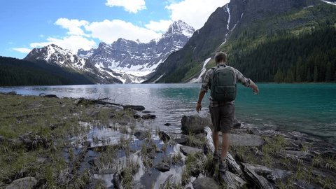 Young man balances on a tree log above stunning mountain lake, man hiker walking on a log by mountain lake in Canada reaches submerged rock and stands arms wide open 