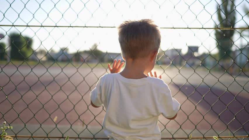 boy stands near the playground. concept of happy childhood and loving family. a child holds on to the net of a sports ground, court and glare of the lifestyle sun in the background